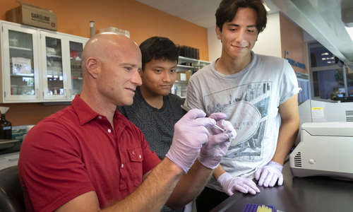 Man in a red shirt holding up something and two students standing behind him watching