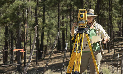 Curator Michele Koons stands behind a large yellow piece of equipment in a wooded area in Reserve, New Mexico