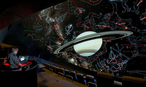 Image of a planetarium screen showing a ringed planet and constellations drawn around it, with a person sitting in the audience controlling the screen.