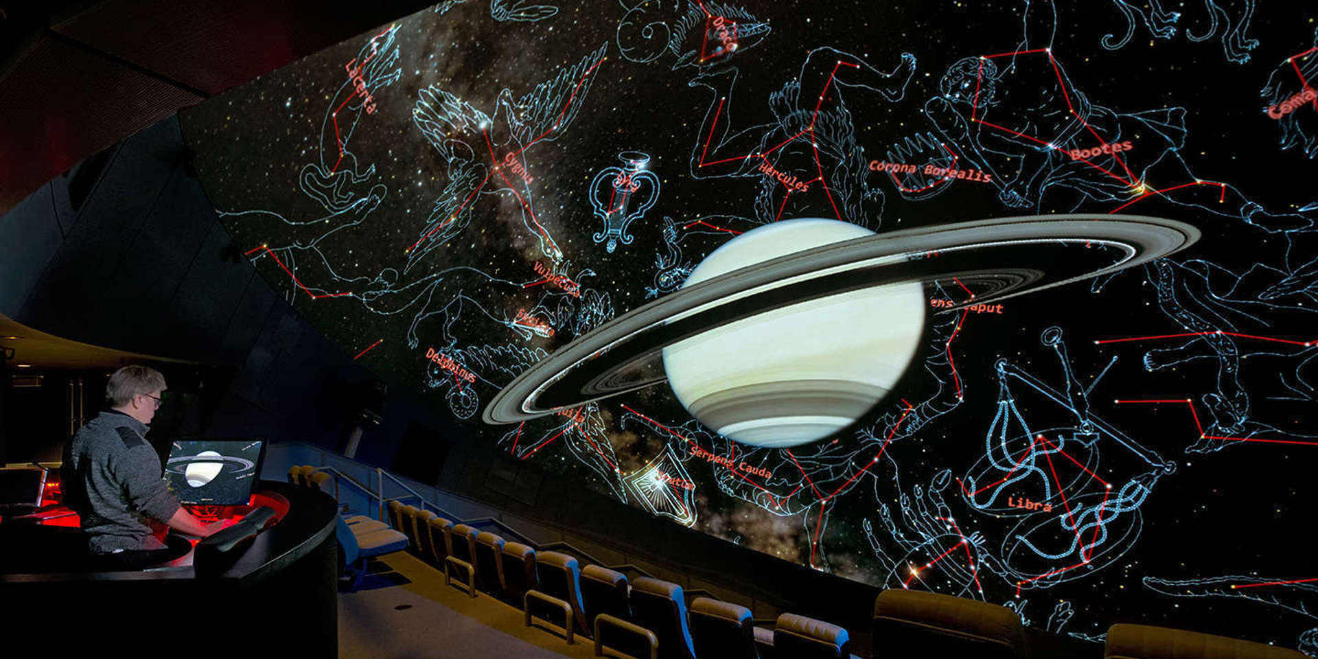 Image of a planetarium screen showing a ringed planet and constellations drawn around it, with a person sitting in the audience controlling the screen.