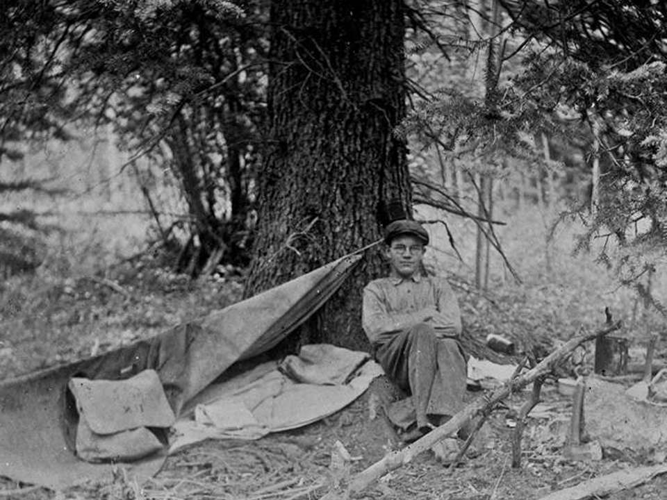 black and white image of a man leaning against a tree with rustic camping equipment around him