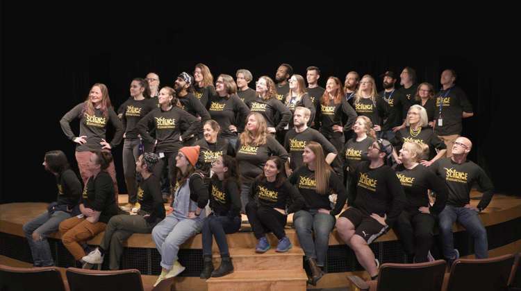 Large group of people wearing the same black shirts standing on a stage all looking to their right.