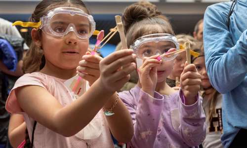 Two girls wearing goggles stretch rubber bands attached to popsicle sticks