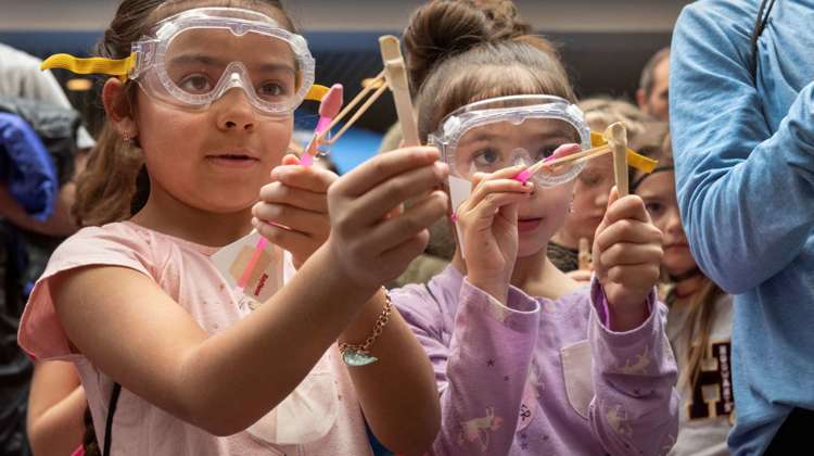 Two girls wearing goggles stretch rubber bands attached to popsicle sticks