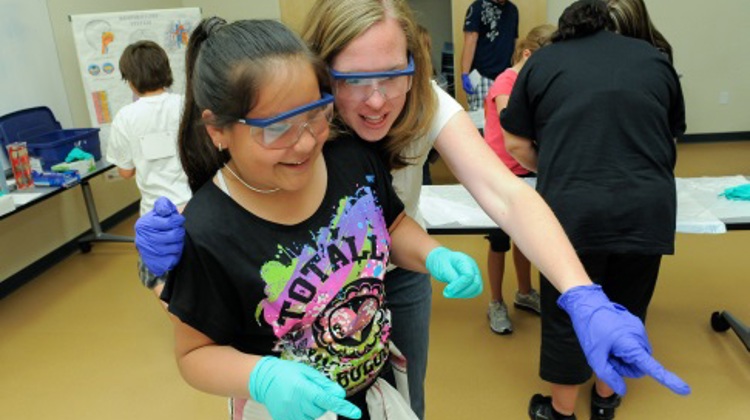 A student and adult wearing gloves smile while pointing at an object out of frame