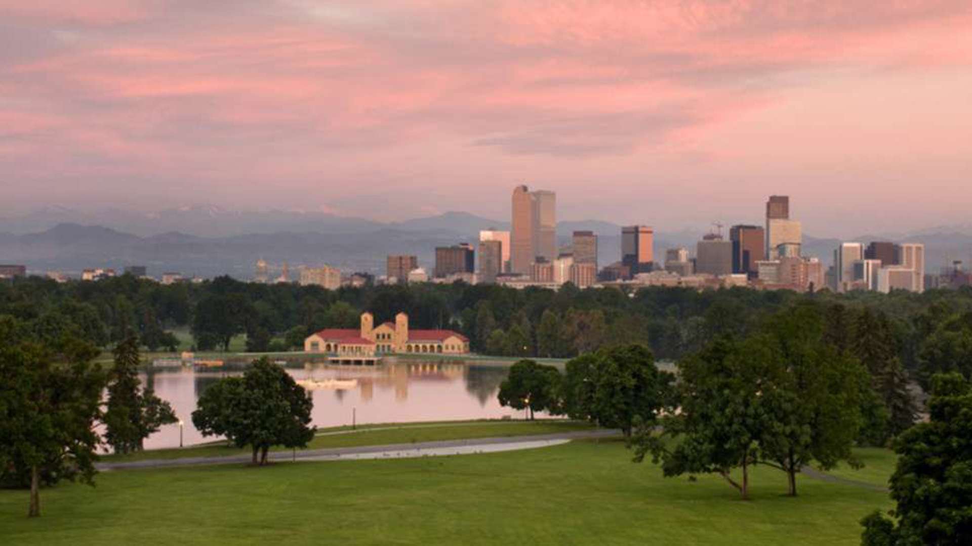 A view of City Park and Downtown Denver including a lake, the skyline, and mountains at sunset