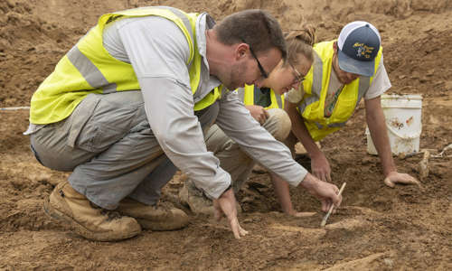 Tyler, Natalie and Salvador are sitting on the ground using tools to inspect a fossil in the ground