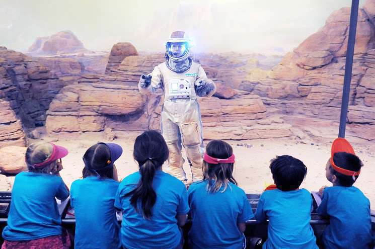 A person dressed in a space suit communicates with children, fostering curiosity about space and encouraging their interest in science.