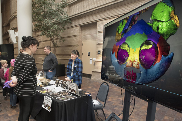 two people standing at a table talking to a guest with a large screen showing digital images