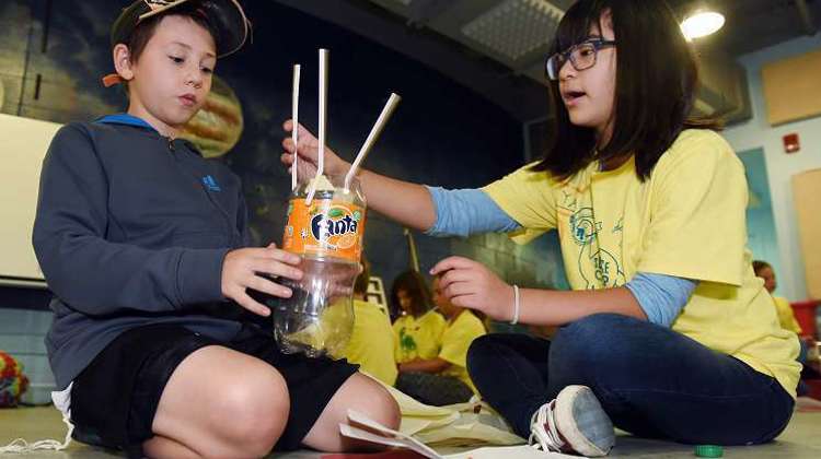 Two students seated on the floor use recyclable materials to create a model