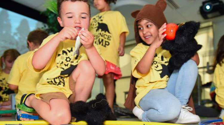 Two children wearing yellow shirts are seated on the floor, happily playing with an assortment of stuffed animals around them.