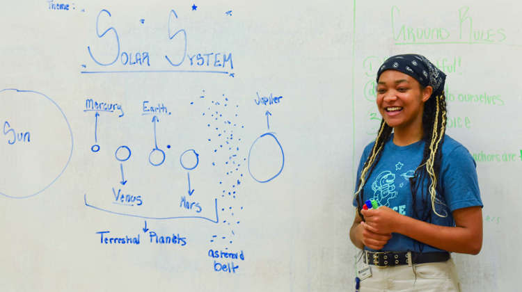 A teacher stands next to a white board with a partial drawing of the solar system.