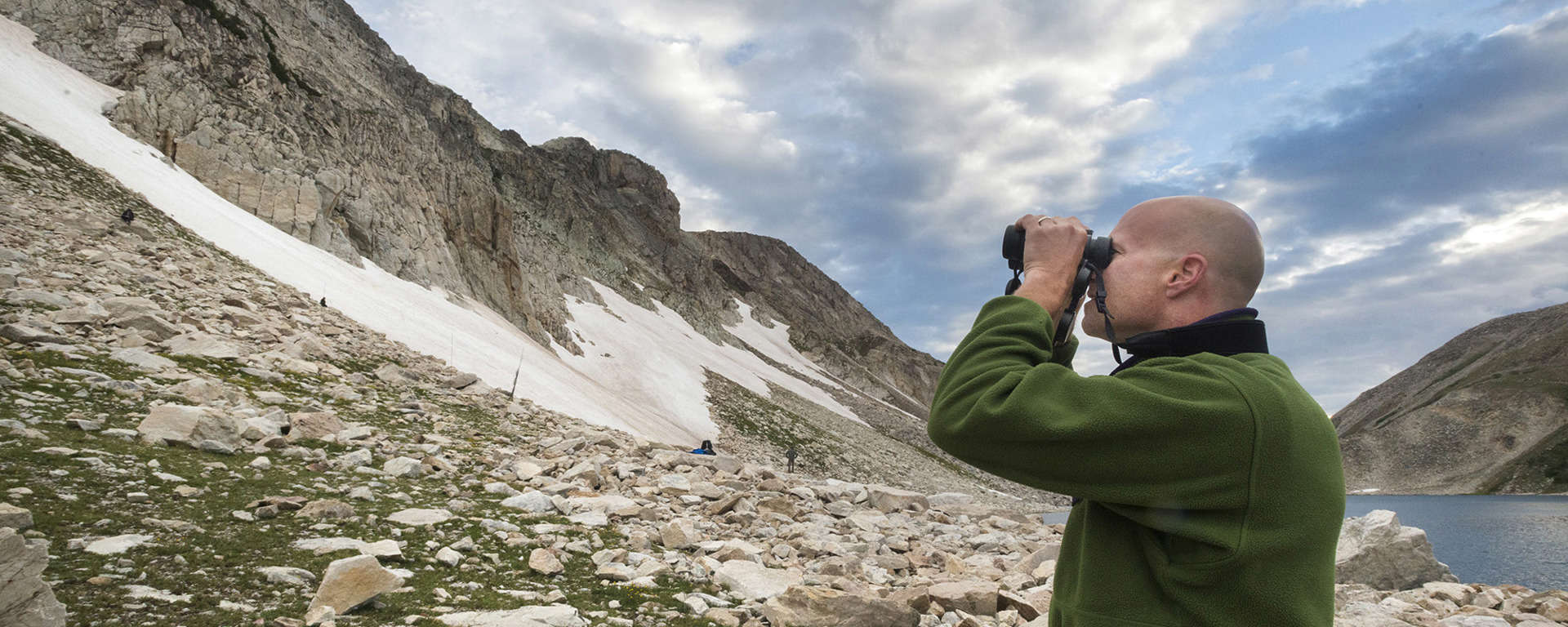 Dr. Garth Spellman stands looking through binoculars at a large snow-covered cliff